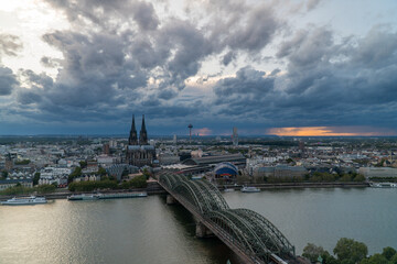 Obraz premium A scenic view of Cologne, Germany, featuring the iconic Cologne Cathedral, the Hohenzollern Bridge, the Rhine River, and a dramatic cloudy sky with hints of sunset light on the horizon.
