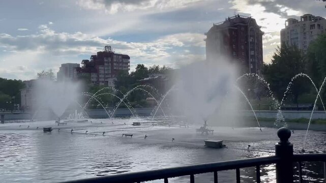 Beautiful fountain spraying water in city pond in summer with green trees around