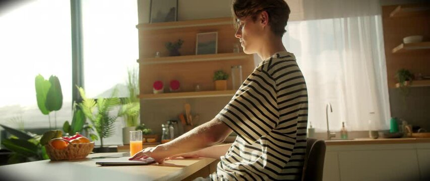 Male Caucasian student in striped T-shirt sitting at table and starting working on laptop. Young man studying online at home. Cozy atmosphere in kitchen. Education concept. Distance learning. Indoors