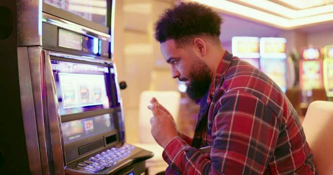 A young man is completely engrossed in the thrilling experience of playing on a colorful slot machine at a lively casino, relishing the excitement of potential victory with every single spin