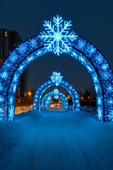 Fototapeta premium Colorful blue snowflake light displays form arches above a snowy pathway, creating a festive atmosphere in a bustling winter city at night