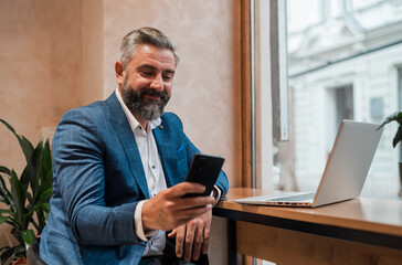 A happy mature business man using his phone while working on a laptop in the office or cafe restaurant.