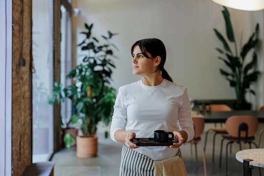 Thoughtful waitress holding tray looking away in cafe