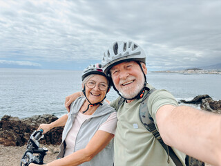 Two happy old mature people enjoying and riding bikes together to be fit and healthy outdoors. Active seniors having fun taking a selfie picture with camera