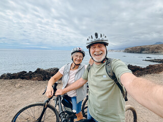 Two happy old mature people enjoying and riding bikes together to be fit and healthy outdoors. Active seniors having fun taking a selfie picture with camera
