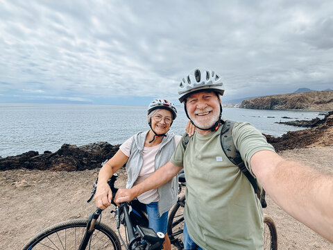 Two happy old mature people enjoying and riding bikes together to be fit and healthy outdoors. Active seniors having fun taking a selfie picture with camera