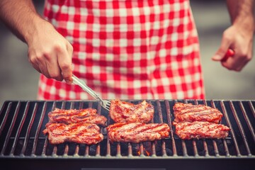 Grilling meat on barbecue grill with red checkered apron by male cook