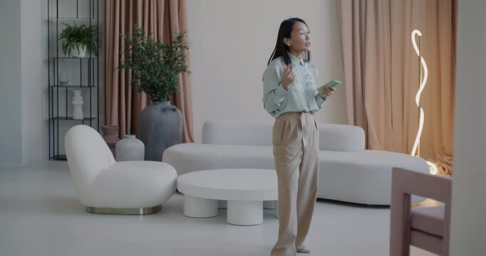 Elegant Asian lady talking on mobile phone through wireless earphones making call walking indoors in luxurious house. Communication and residence concept.