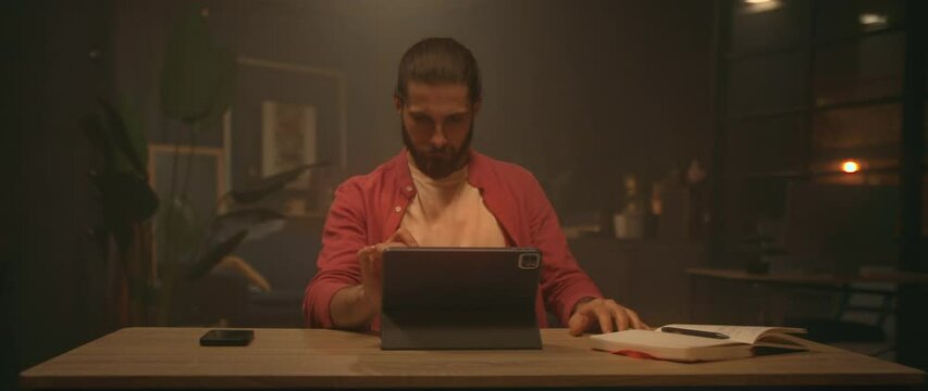 Shot of inspired handsome young Caucasian man sitting at desk and using digital tablet for drawing image of woman. Guy using stylus. Modern living room. Remote work concept. Freelancer routine