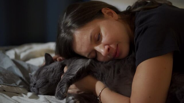 Adult 44 year old woman and gray domestic cat lying on bed in bedroom. Happy woman hugging and kissing her cat, stroking his belly.