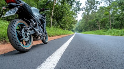 Fototapeta premium Close-up view of a black motorcycle's front wheel and headlight on a quiet asphalt road surrounded by lush green trees in soft light