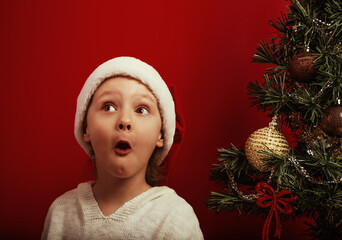 Emotional kid girl with surprising eyes and open excited mouth grimacing near the decor holiday fur green tree in red santa claus hat. Happy expression winter holadays with natural emotion child