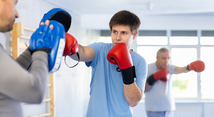 Focused adolescent guy wearing boxing gloves working out with male coach during self-defense...