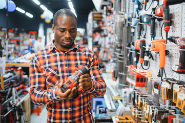 Fototapeta premium African american man holding and choosing electric screwdriver, standing in construction store