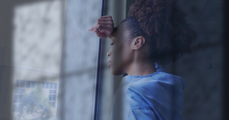 Image of rain on window over unhappy african american female doctor looking out of window