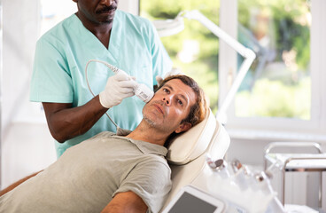 Obraz premium Middle-aged male patient experiencing facial cryotherapy procedure using ice hammer to tighten skin in aesthetic medicine clinic. Advanced hardware cosmetology techniques