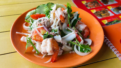 Spicy seafood salad with shrimp, squid, and pork, presented on a bright orange plate, alongside a menu, highlighting a delicious dining experience
