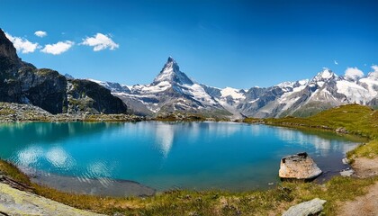 Naklejka premium Panorama of the Alps: Clear mountain lake with Matterhorn in the background, surrounded by lush greenery and snow-capped peaks under a blue sky