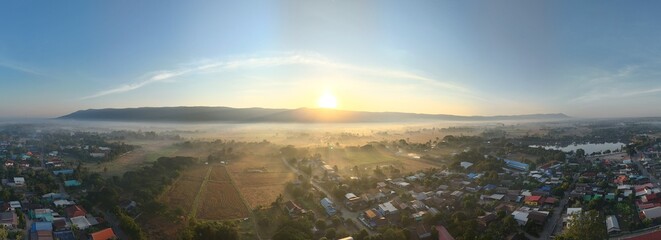 Panoramic aerial view of a tranquil rural village at sunrise, with golden sunlight streaming through the morning mist. 