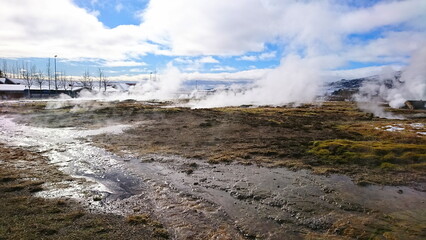 Haukadalur, Iceland - 3.24.2018: Steam coming out from geysers among the grassland under a cloudy blue sky with a visitor centre and restaurants before the pandemic 