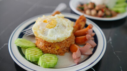 Plate of Thai-style fried rice topped with a sunny-side-up egg served with sliced sausages, crispy fried chicken and fresh cucumber slices.