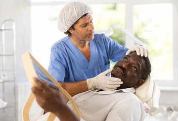 Experienced male cosmetologist examines facial skin of African-American man patient and advises on...