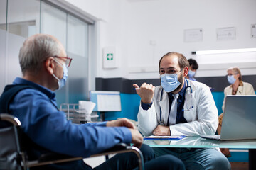 Young doctor discussing recovery plans with retired man in wheelchair, having laptop nearby on desk for access to medical records. Pensioner patient listening to medic specialist during consultation.