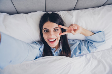 Charming young woman enjoying a fun morning in bed with a peace sign and a cheerful smile in cozy blue pajamas