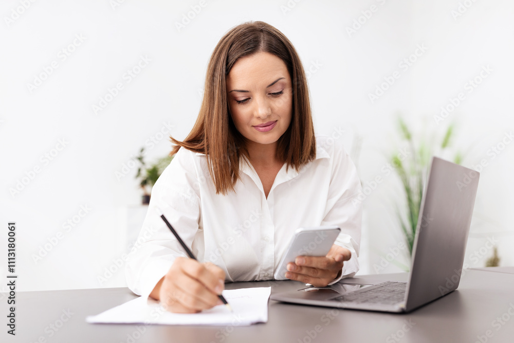 Poster A businesswoman actively engages in her work at a modern office. She uses a smartphone to check information while taking notes on a notepad, immersed in her tasks.
