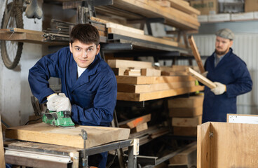 Young guy worker in carpentry workshop polishes wooden board with machine