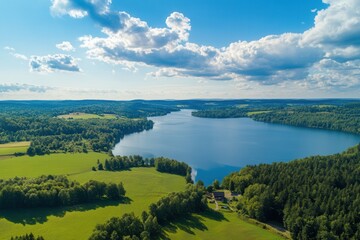 Obraz premium Aerial view of serene lake surrounded by lush green fields and forests under a vibrant blue sky with fluffy clouds.