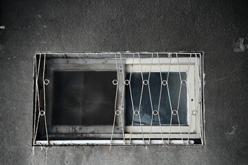 An aging cement wall bears black dust, framing a white-painted wooden window with iron gratings....
