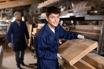 Young guy worker takes wooden plank from stack in wood workshop