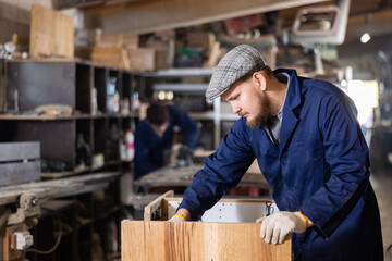 Young male carpenter assembles wooden cabinet from parts in wood workshop