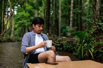 young asian man enjoying sitting on wooden chair drinking coffee together in middle of a stream forest.