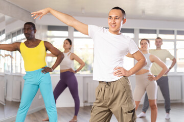 Cheerful Asian guy in casual sportswear participating in group Latin dance lesson, gracefully performing salsa steps in spacious well-lit studio