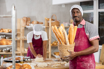Smiling successful African American baker holding rustic wicker basket of golden crispy baguettes,...