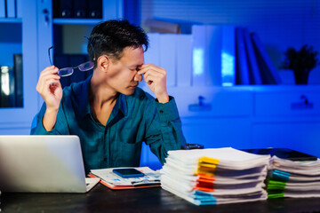 Fototapeta premium An Asian businessman works late at night in the office,dealing with stress,migraine headaches,overtime.Checking documents,experiencing office syndrome symptoms,fatigue,insomnia risks from prolonged