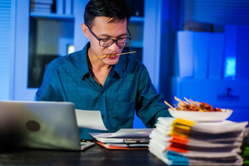 An Asian young businessman sits at his desk working late at night. Hungry and busy, he eats mala from a plate while checking and managing numerous documents under time pressure