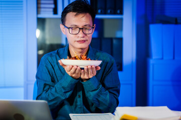 An Asian young businessman sits at his desk working late at night. Hungry and busy, he eats mala...