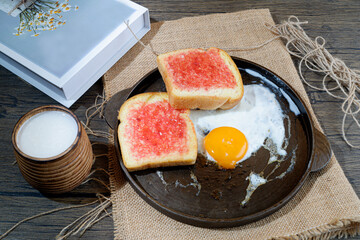 A rustic breakfast spread featuring a fried egg on a cast iron skillet, two slices of toast with strawberry jam, and a glass of milk.