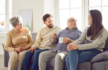 Happy cheerful smiling senior old parents and their adult children man and woman spending time together sitting on sofa in living room at home and talking with cup of tea. Family leisure concept.