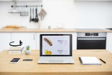 Laptop on wooden desk in modern kitchen showing fast food online sales site with pricing and discounts. Desk includes smartphone, credit card, notebook, pen, and headphones.