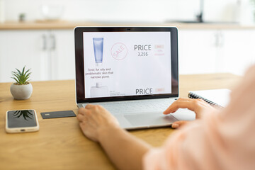 Young Caucasian woman shops online for skincare products using laptop at home. She reviews a website for cosmetics specializing in oily skin, with a focus on toner sale.