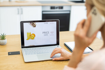 Caucasian woman in kitchen uses laptop to browse online food delivery while on phone. Screen displays food prices and discounts. Credit card and notebook on table indicate online shopping and planning