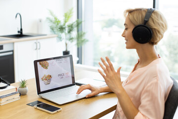 Happy Caucasian woman ordering food online using laptop while listening to music in headphones. She sits at desk in modern room, engaged in digital shopping experience.