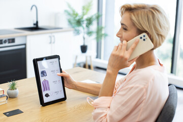 Caucasian woman in her 30s using phone and tablet to shop online in modern home office. She browses clothing items on tablet screen while engaged in phone call, surrounded by contemporary decor.