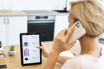 Caucasian woman using tablet and smartphone to shop for clothes online. She is browsing online delivery website, placing order for fashion items. Image focuses on modern technology in everyday life.
