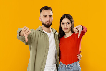 A young Caucasian couple stands close together, making thumbs down gestures. Their expressions convey frustration or disapproval, set against a bright yellow background.