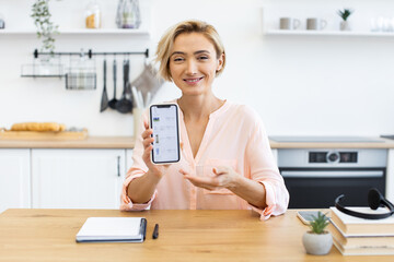 Happy Caucasian woman in casual attire holding smartphone showing online cosmetics delivery app. Sitting in modern kitchen, she displays product shopping service.